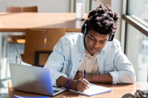 An image of a male student wearing headphones writing notes at a desk.